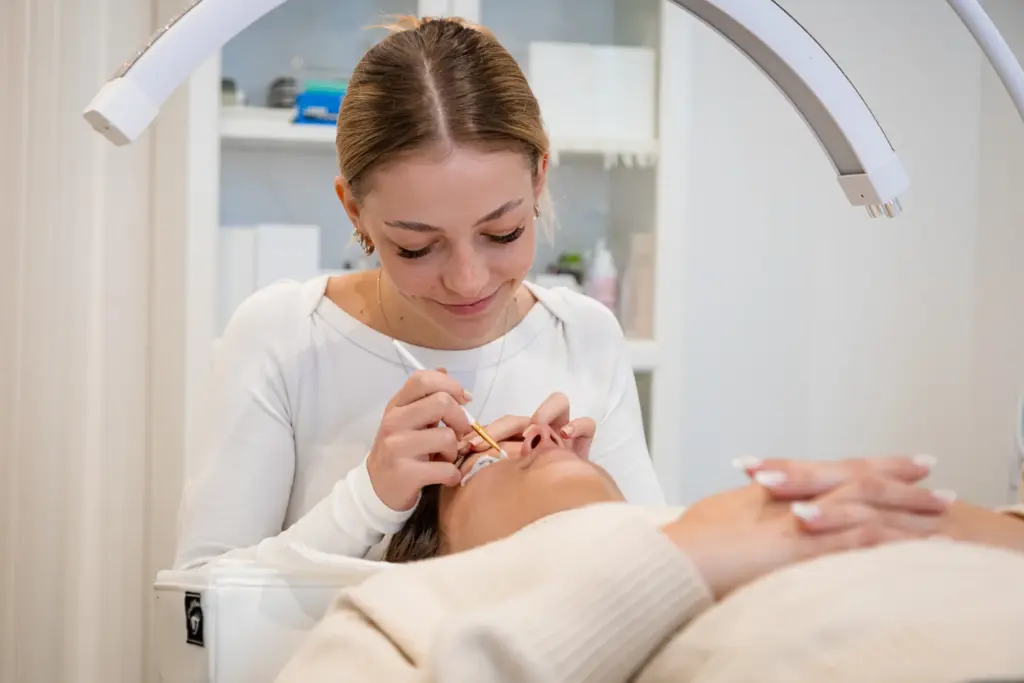 Szempillastylist precíz eszközzel dolgozik egy kezelőágyon fekvő vendég szempilláin.