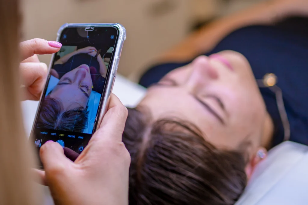 Pink körmös kéz telefonnal fotózza a fekvő nő arcát és szempilláit.