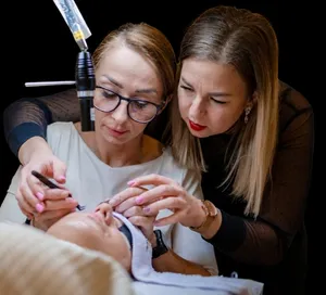 Két nő szempilla-stylingot gyakorol egy fekvő vendégen, nagyító lámpa fényénél.