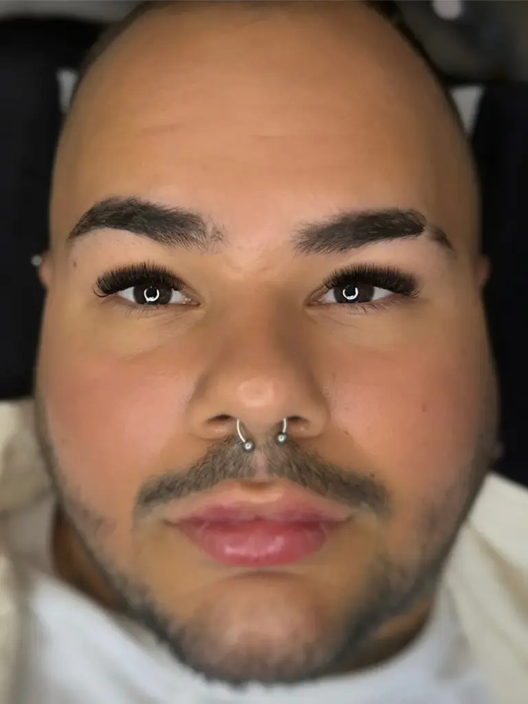 Közelkép egy arcról, dús műszempillákkal, formázott szemöldökkel és septum piercinggel.